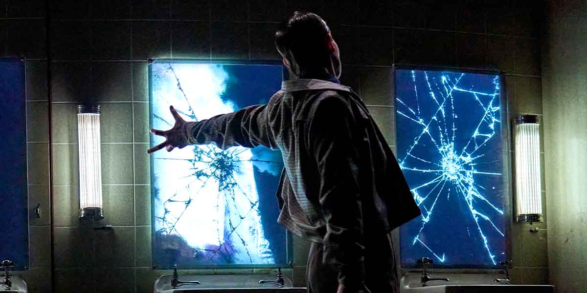 A person in a checked shirt reaches towards a cracked, illuminated mirror in a dimly lit bathroom.