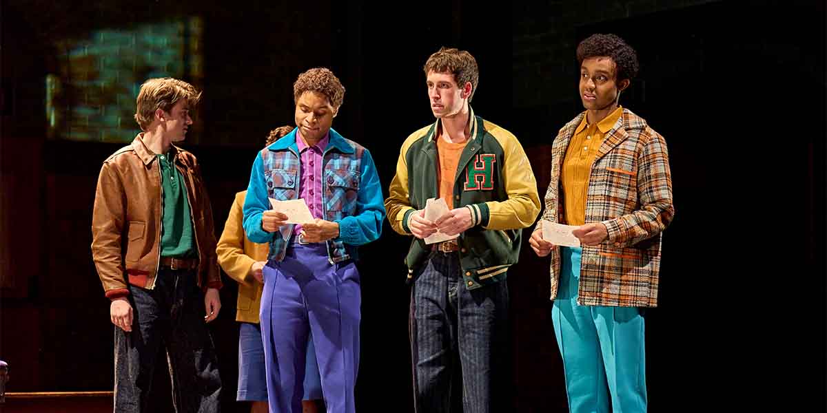 Four young men stand together on stage, holding papers, dressed in colourful 1980s-inspired outfits.
