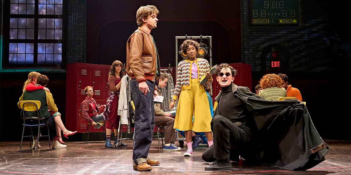A group of young actors performs on stage, showcasing vibrant costumes and dynamic poses in a school setting.