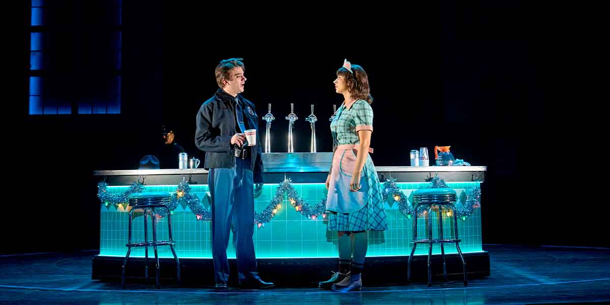 A man and woman converse at a brightly lit bar, decorated with festive garlands, in a theatrical setting.