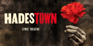A scene from Hadestown featuring performers in period costumes, with warm lighting and a dramatic backdrop.