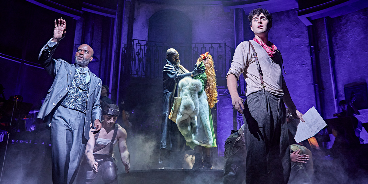 Four performers in theatrical costumes, standing together on stage, with dramatic lighting and a dark background.