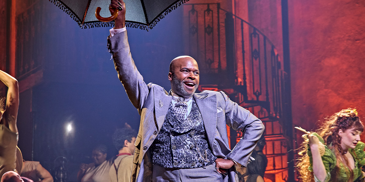 Cedric Neal performs on stage in a vibrant costume, surrounded by dramatic lighting and theatrical set pieces.