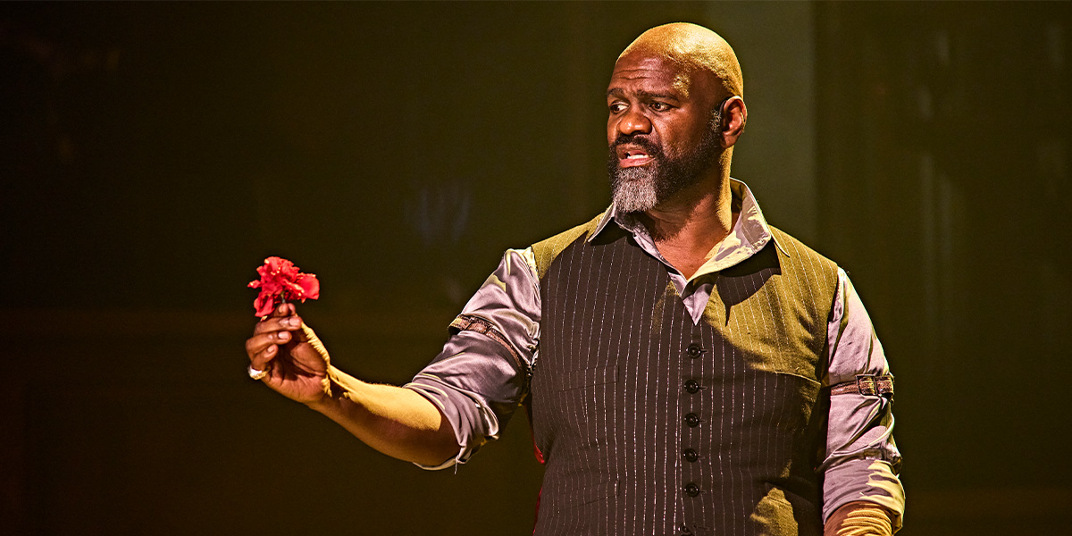 Chris Jarman as a character in Hadestown, wearing a dark costume, performing on a dimly lit stage.