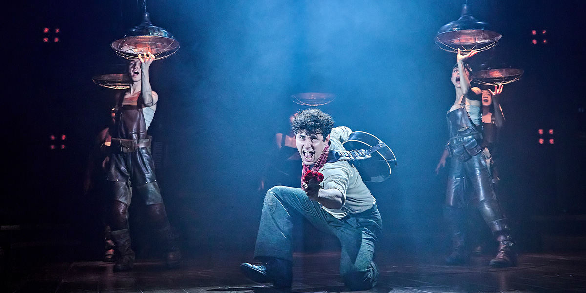 Dylan Wood performs on stage in Hadestown, wearing a dark costume, with dramatic lighting and set design.