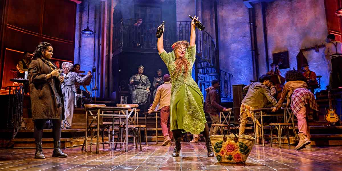 Fayth Ifil and Danielle de Niese perform on stage with the cast of Hadestown, vibrant costumes visible.