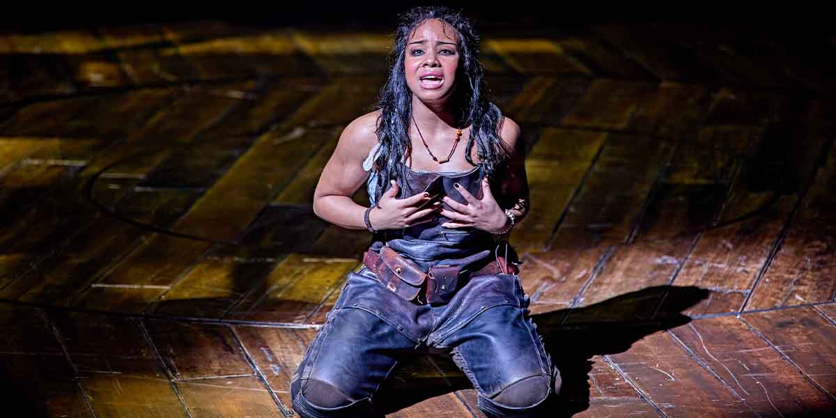 Fayth Ifil kneels on a wooden stage, expressing emotion with her hands on her chest.