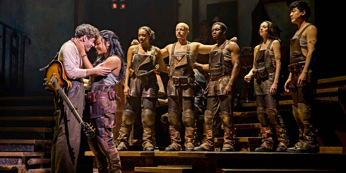 Nathan Sykes and Fayth Ifil embrace, surrounded by cast members in brown costumes on a stage.