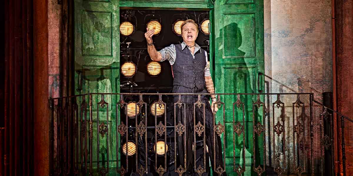 Shaun Dooley performing in Hadestown, wearing a dark costume, with dramatic lighting and a theatrical backdrop.