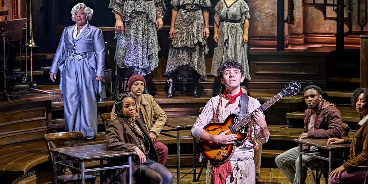 The cast of Hadestown poses together, wearing period costumes in earthy tones, with a theatrical backdrop.