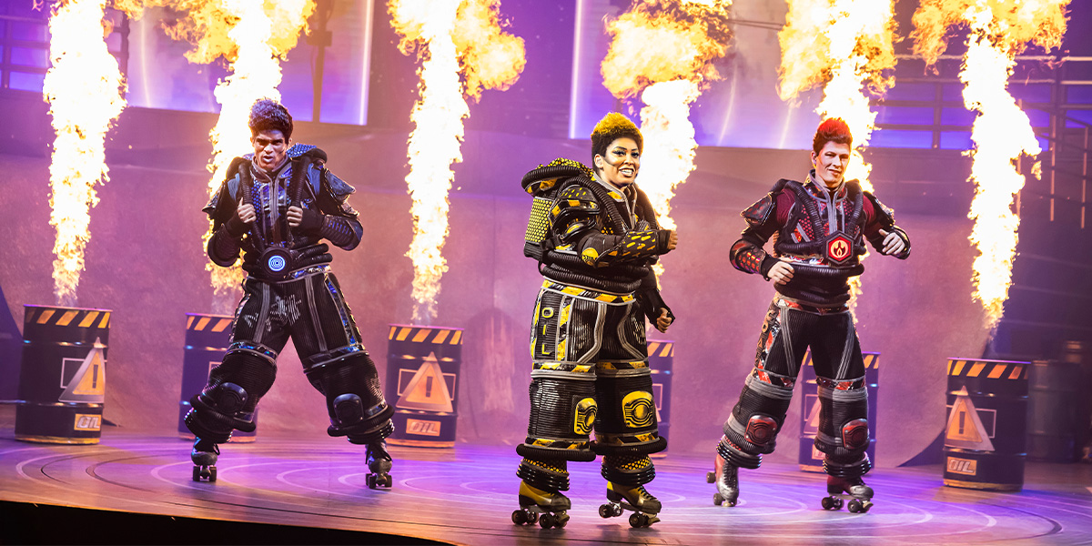 A performer in a vibrant costume strikes a dynamic pose on stage, with colourful lights in the background.