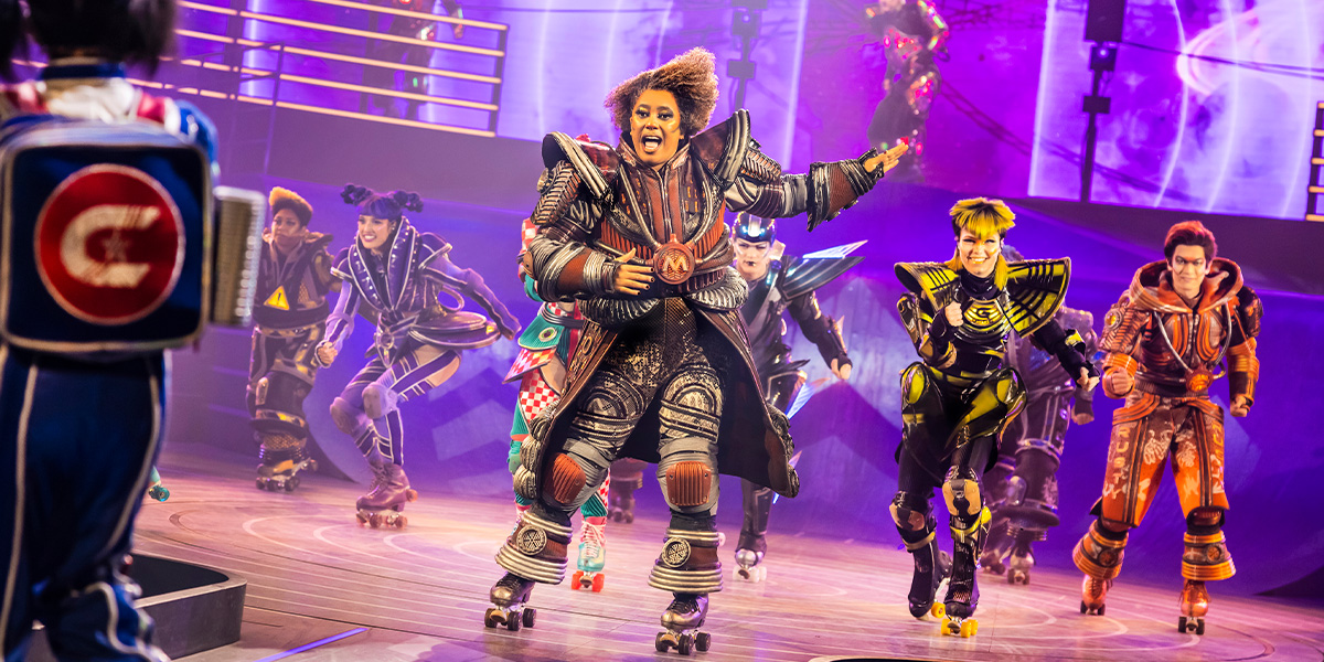 A performer in a vibrant costume strikes a dynamic pose on stage, illuminated by colourful stage lights.