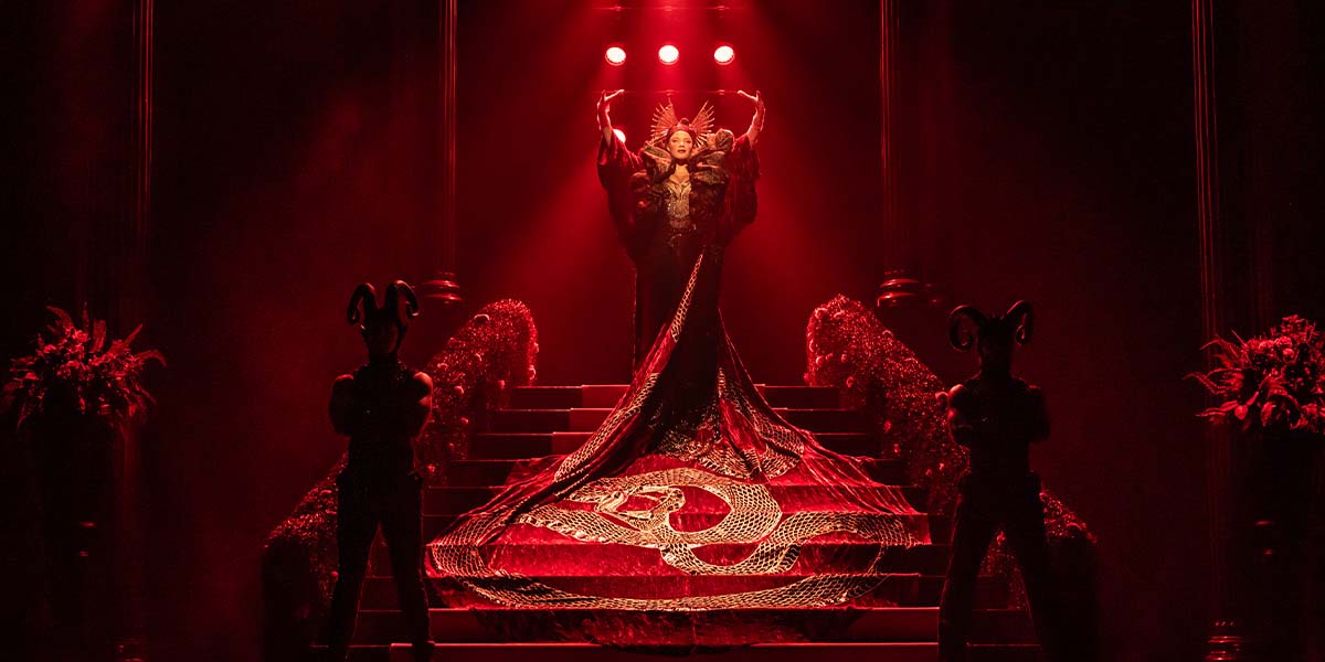 A dramatic scene featuring a figure in a red gown with intricate patterns, flanked by two dark figures.