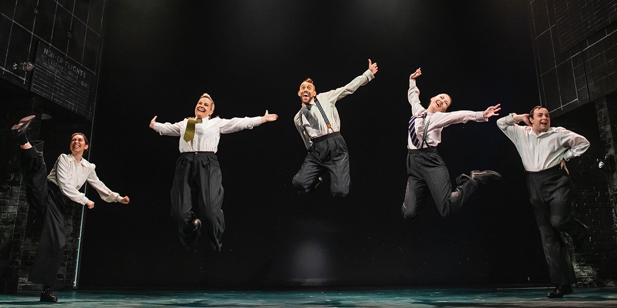 Five performers in formal attire joyfully jumping against a dark stage backdrop.