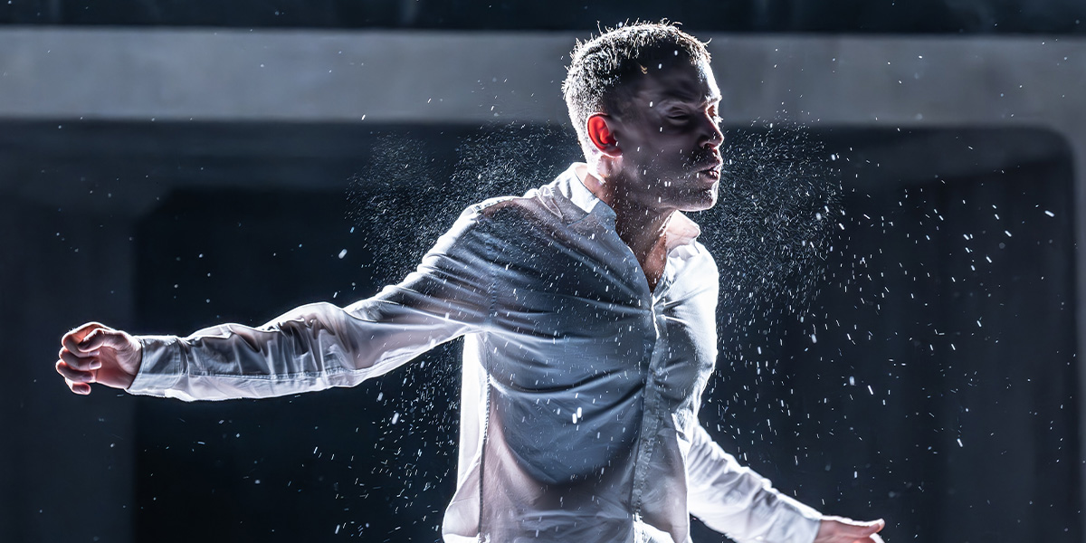 David Shields as Jacob Dunne, wearing a dark suit, gestures expressively against a dimly lit stage backdrop.