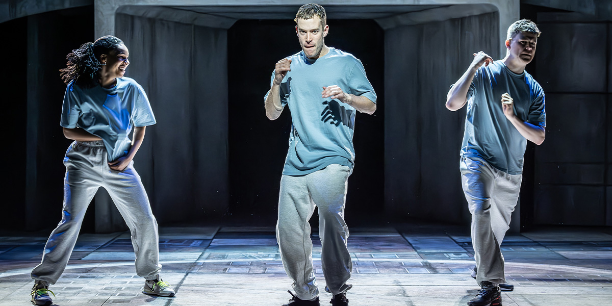 Three actors on stage, engaged in a dramatic scene, with varied expressions and costumes against a dimly lit background.