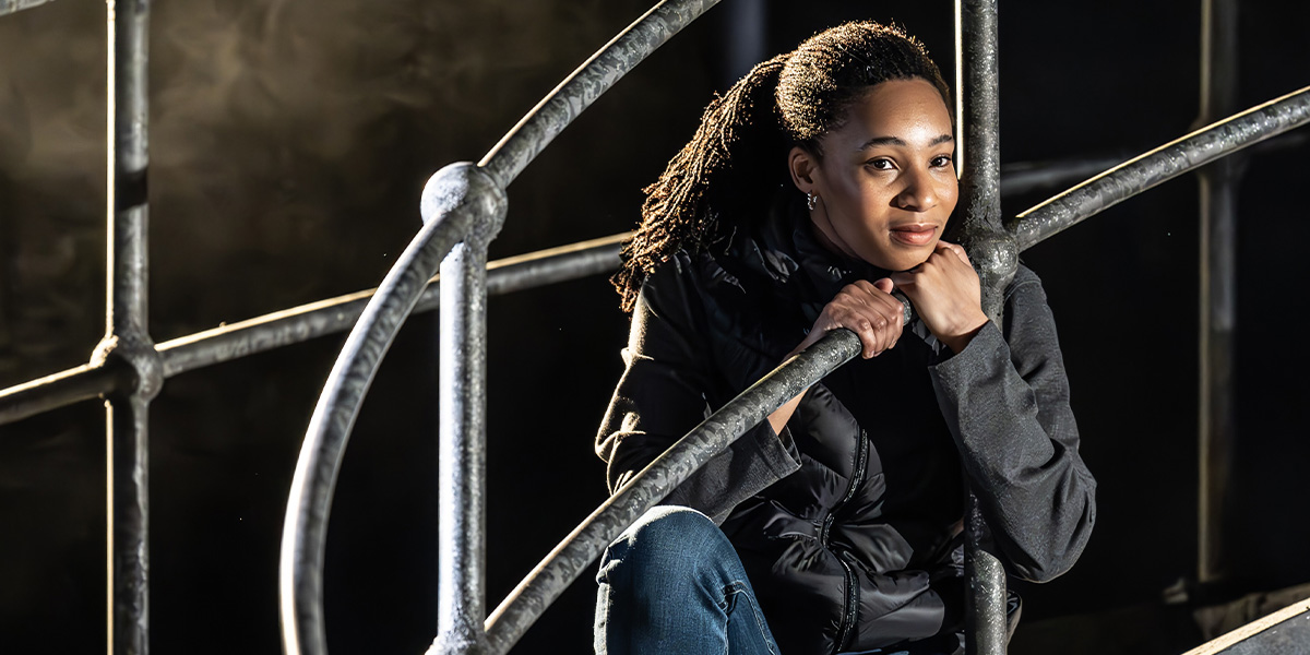 Shalisha James-Davis performs energetically on stage, wearing a vibrant costume, with dramatic lighting in the background.