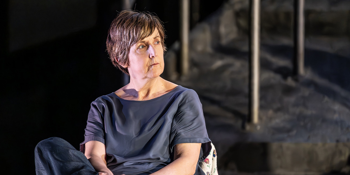 Julie Hesmondhalgh performs on stage, wearing a vibrant costume, with dramatic lighting and a textured backdrop.