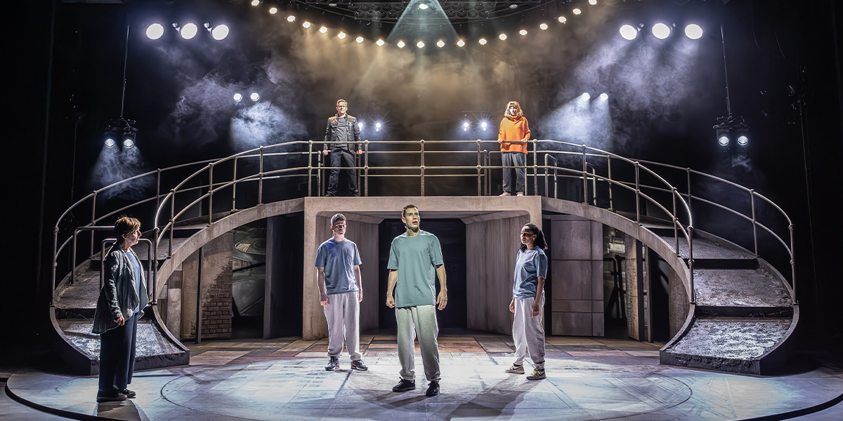 David Shields stands centre stage with the cast of Punch, dressed in colourful costumes, performing energetically.