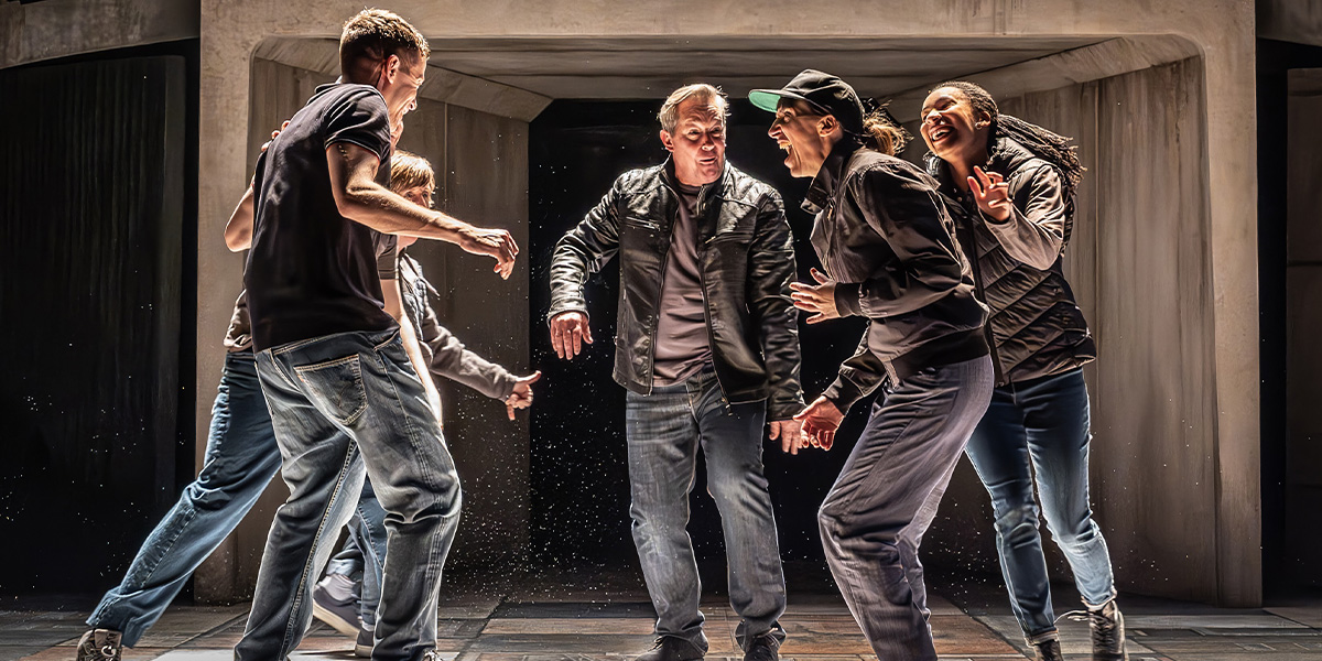 Tony Hirst stands centre stage, surrounded by puppets and cast members, under warm stage lighting.