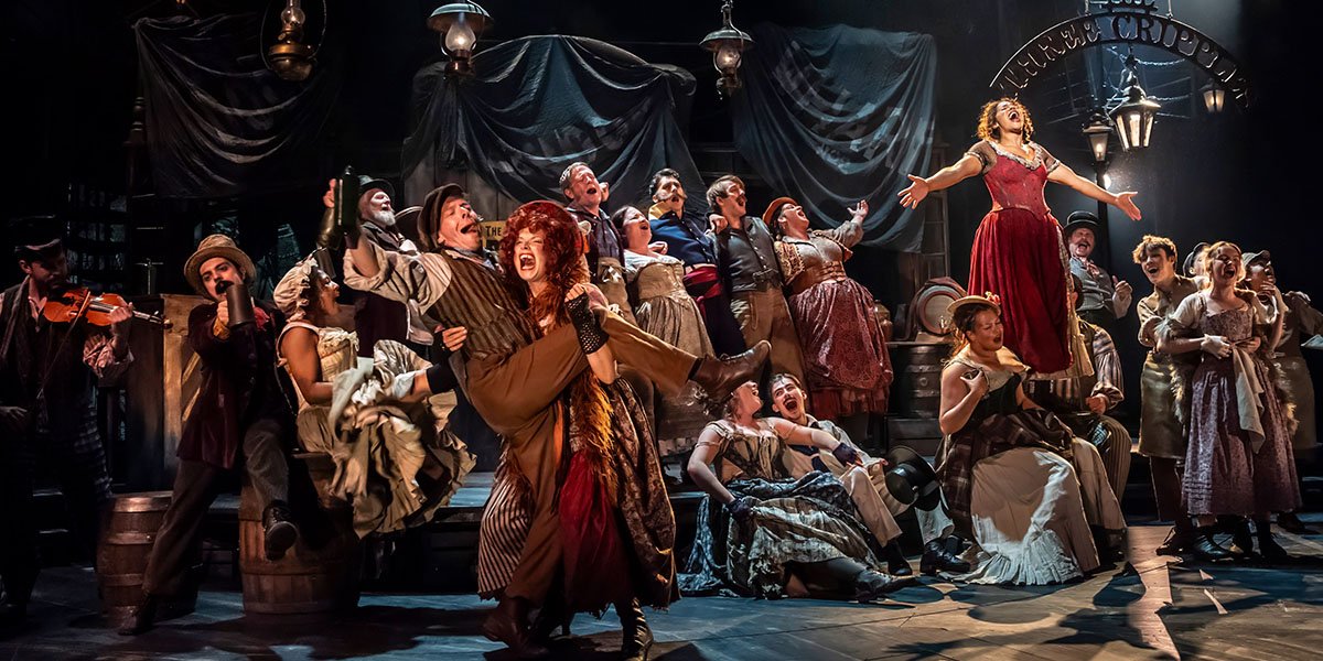 Oliver stands centre stage, wearing a bright red costume, with a vibrant backdrop and dramatic lighting.