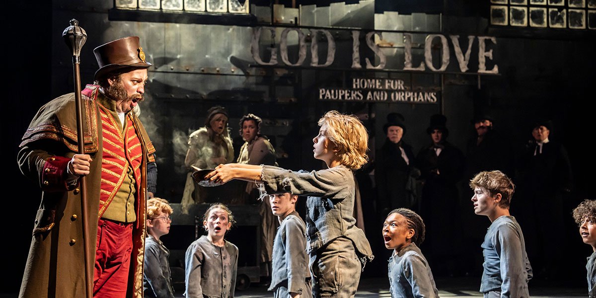 Oliver stands centre stage, wearing a vibrant costume, with dramatic lighting and a cheering audience in the background.