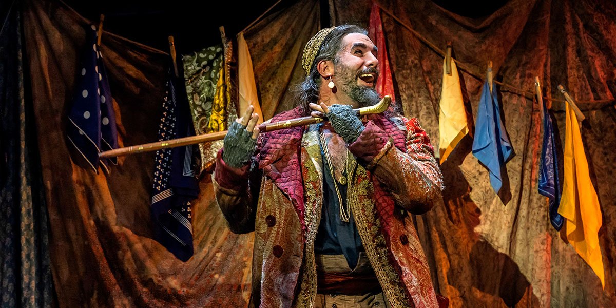 A man in a black suit and white shirt stands confidently, smiling against a blurred stage backdrop.