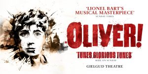 Oliver stands centre stage in a theatrical pose, wearing a tattered brown coat, with a spotlight overhead.