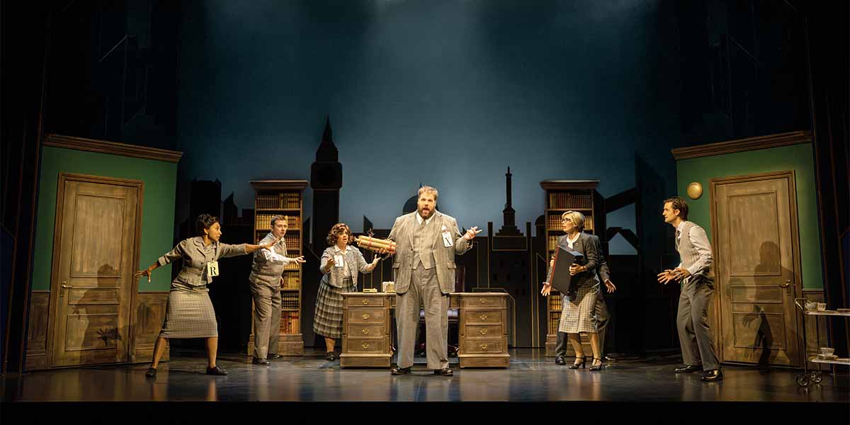 A group of six actors in vintage costumes perform on stage, surrounded by wooden furniture and a cityscape backdrop.