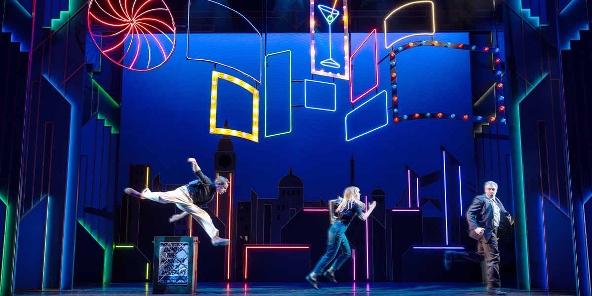 Three performers are on stage, with vibrant neon lights and a city skyline backdrop.