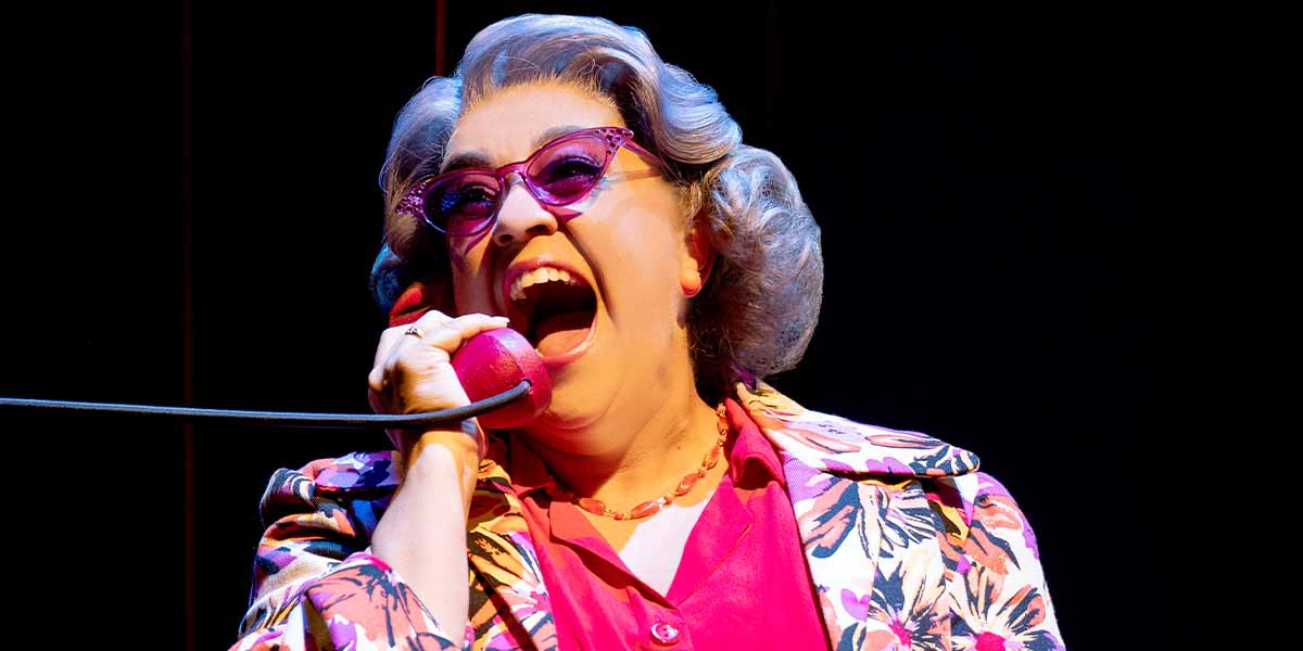 Nancy Zamit passionately performs on stage, holding a red telephone, wearing a floral jacket and glasses.