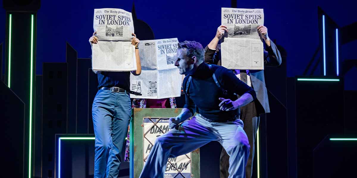 Three performers hold newspapers titled "SOVIET SPIES IN LONDON," while one acts dramatically in front.