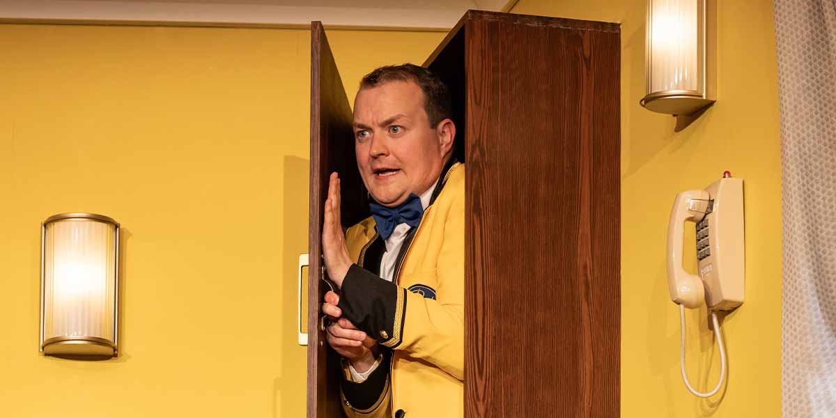 Greg Tannahill, wearing a yellow jacket, emerges from a wooden cupboard against a yellow wall.