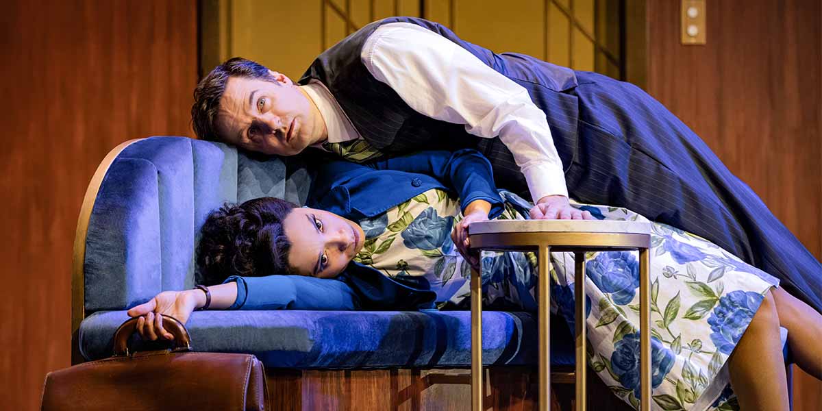 Henry Shields leans over Adele James, who reclines on a blue sofa, surrounded by a wooden backdrop.