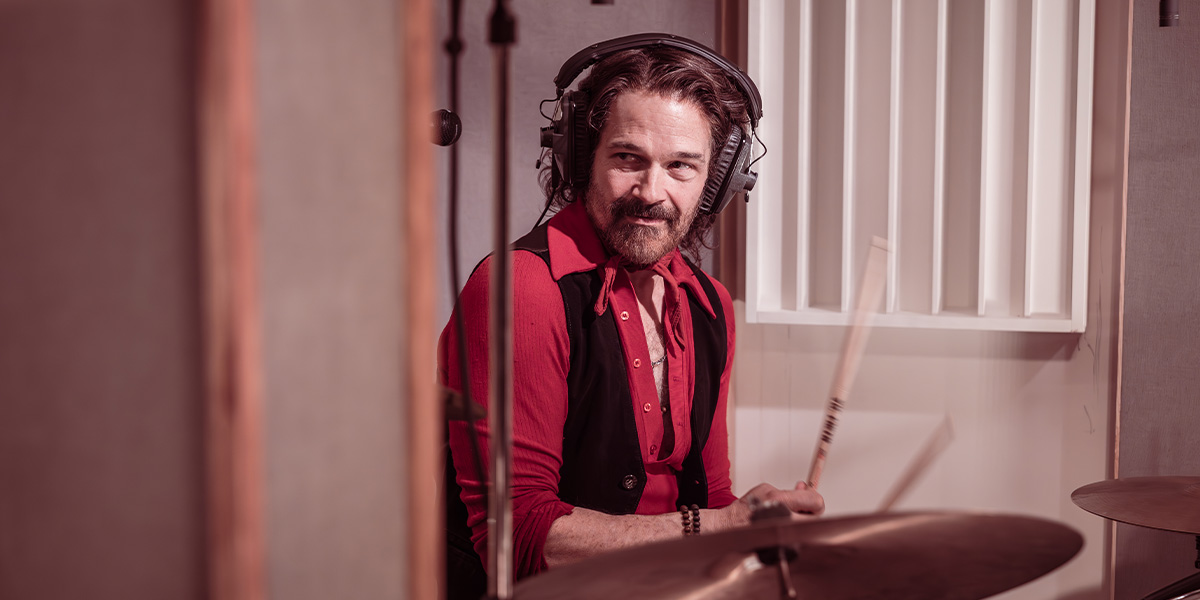 A musician plays guitar on stage, illuminated by colourful lights, with a cheering audience in the background.