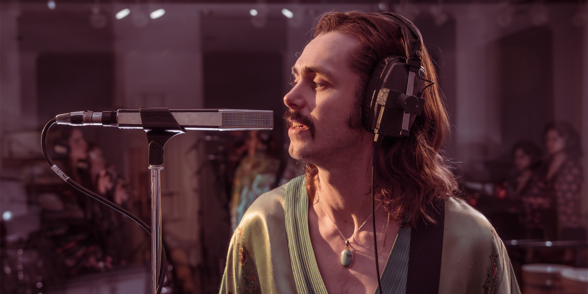 A musician plays a guitar on stage, illuminated by colourful lights, with a blurred audience in the background.