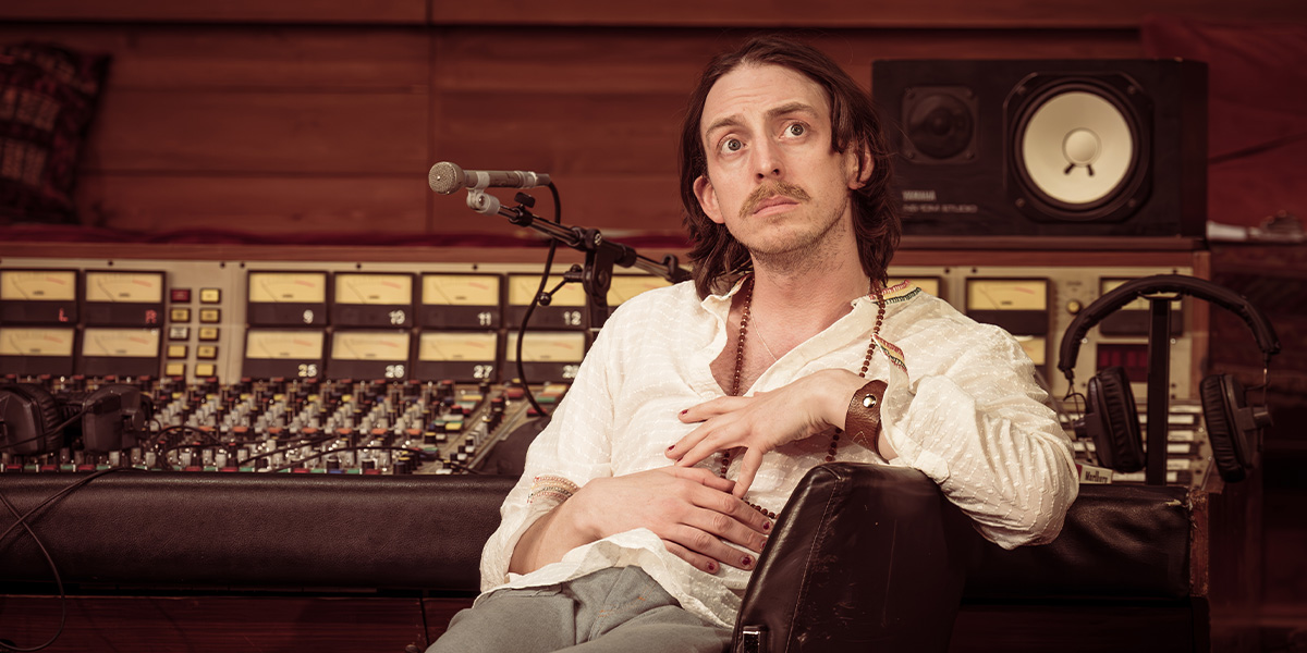A musician plays a guitar on stage, wearing a black shirt, with colourful stage lights in the background.