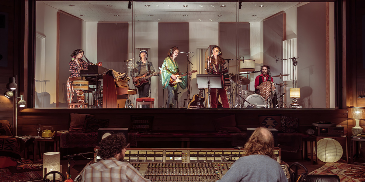 A band consisting of five musicians performs in a recording studio equipped with various instruments including keyboards, guitars, and drums. The scene includes a person at a mixing console, overseeing the recording session.