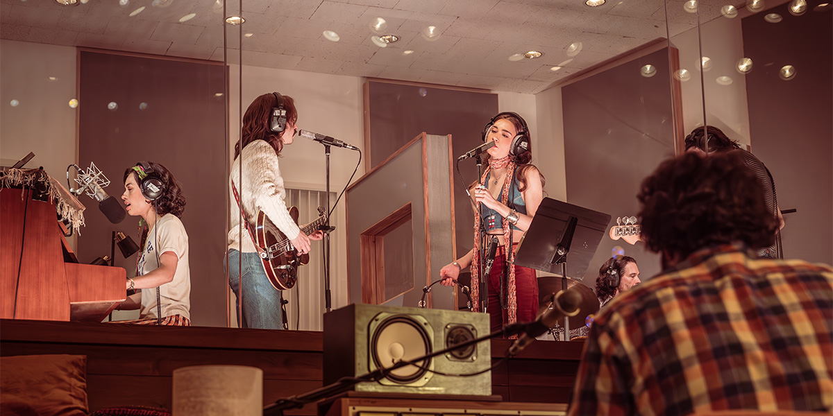 A musician plays a guitar on stage, illuminated by colourful lights, with a cheering audience in the background.