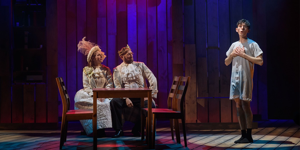 On the left two people who are regally dressed sit at a table. On the right stands a person wearing a white all in one pyjamas.