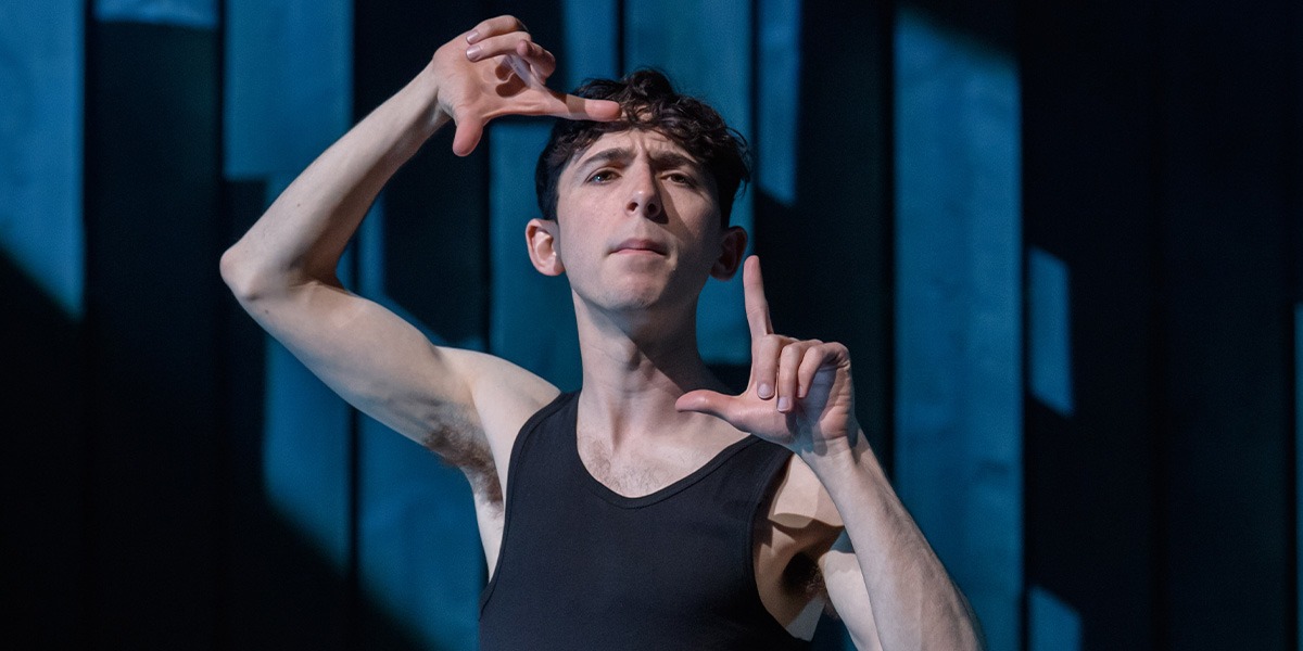 A person is standing in front of a blue background making a gesture with their hands which suggests taking a snapshot.