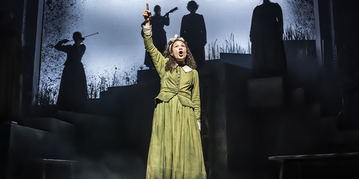 Allyson Ava-Brown as Nell, wearing a dark costume, stands centre stage with dramatic lighting in Coven.