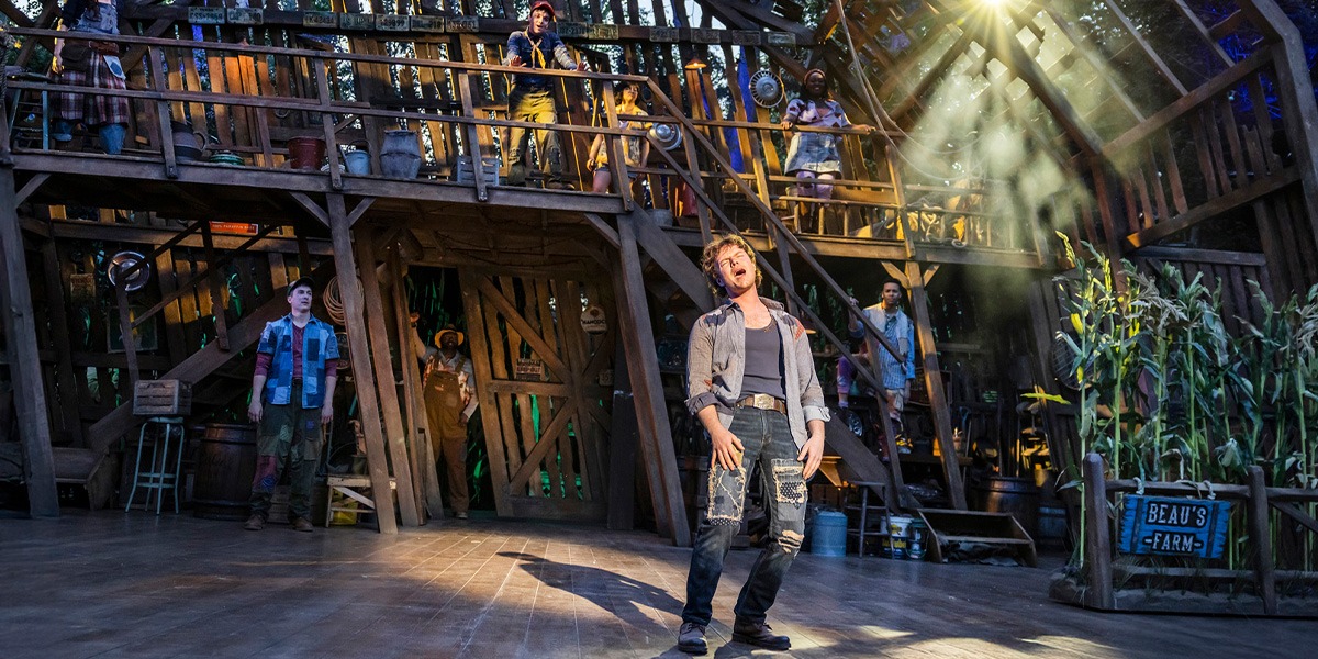 Ben Joyce (Beau) is standing in the spotlight, centre stage, surrounded by other company members in a rustic barn-like set, enhanced with theatrical lighting.