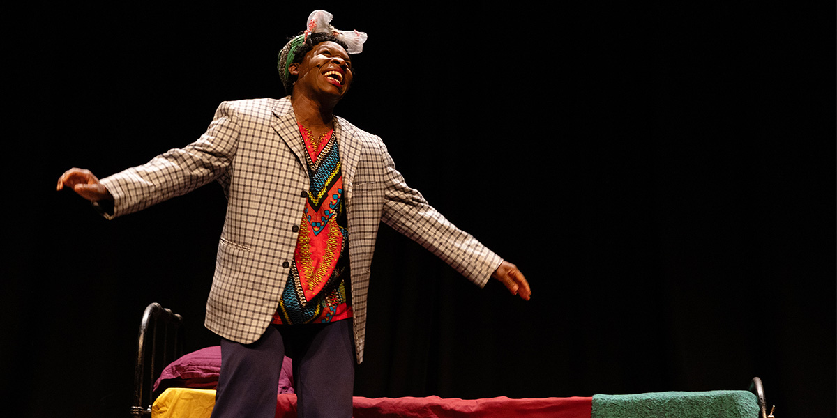 Apphia Campbell performs joyfully in a colourful outfit, standing beside a bed against a dark background.