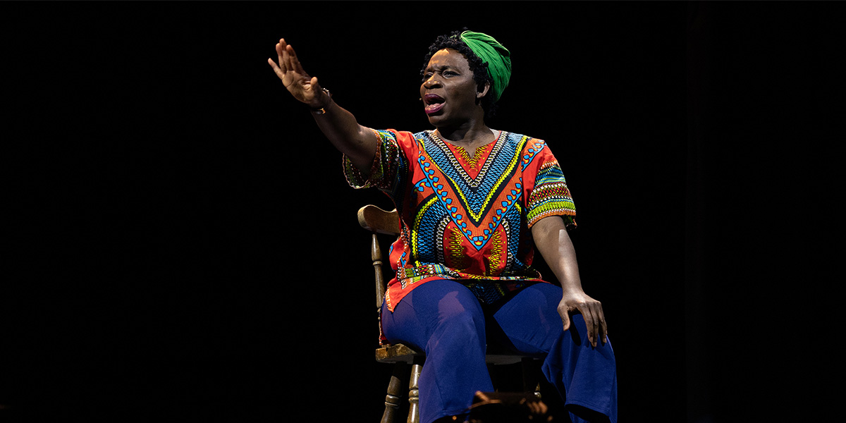Apphia Campbell performs passionately on stage, wearing a vibrant patterned top and blue trousers, against a dark background.