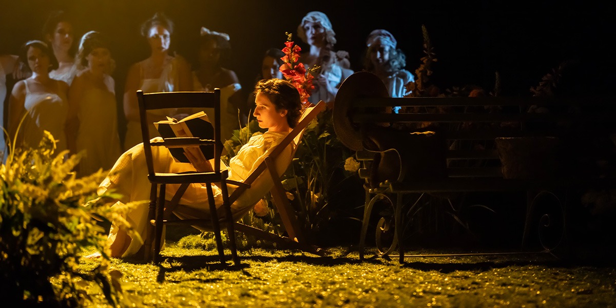 Bessie Carter (Vivie Warren) is sitting in a deck chair, surrounded by wild flowers and reading a book.