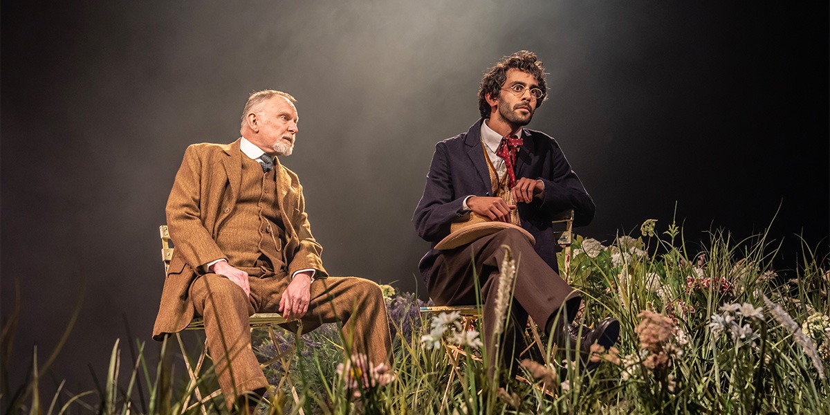 Robert Glenister (Sir George Crofts) and Sid Sagar (Mr. Praed) are sitting on wooden chairs surrounded by wild flowers.