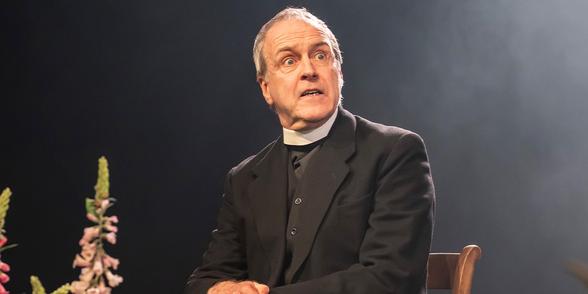 Kevin Doyle (Rev. Samuel Gardner) is sitting on a wooden chair and looks suprised.