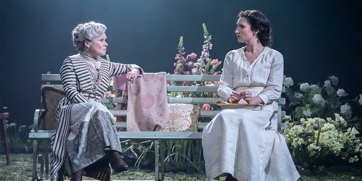 Bessie Carter (Vivie Warren) and Imelda Staunton (Mrs. Kitty Warren) sit on a wooden bench facing one and other, both appear to be unhappy.
