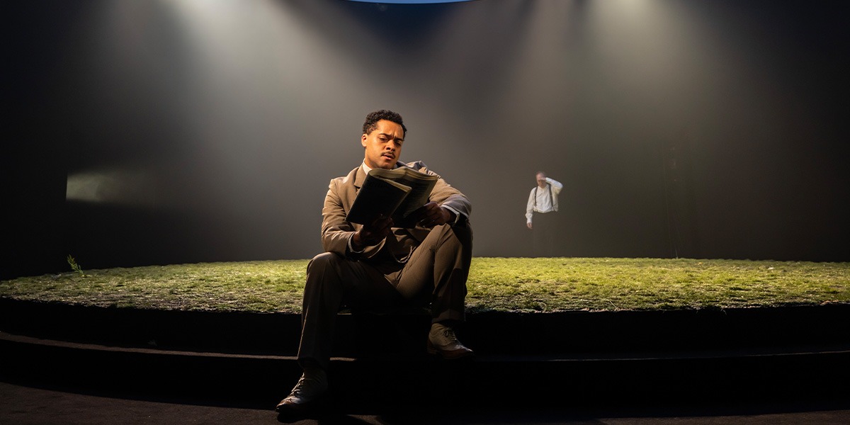 Reuben Joseph (Frank Gardner) is sitting on green grass and is reading a newspaper.